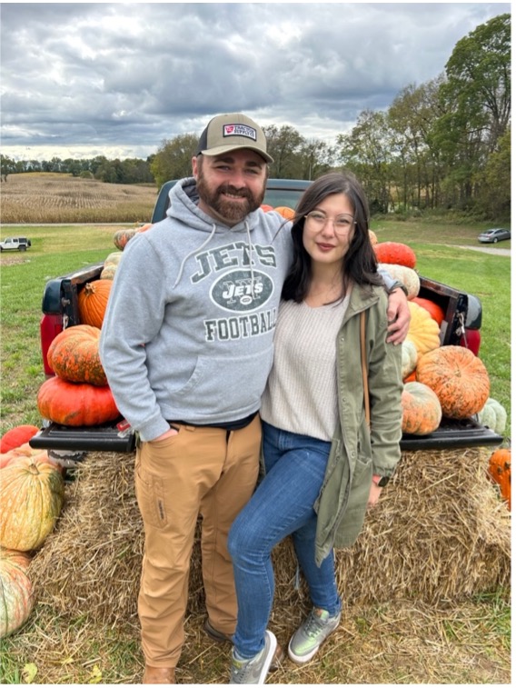 Two people standing in front of a truck full of pumpkins and hay stacks