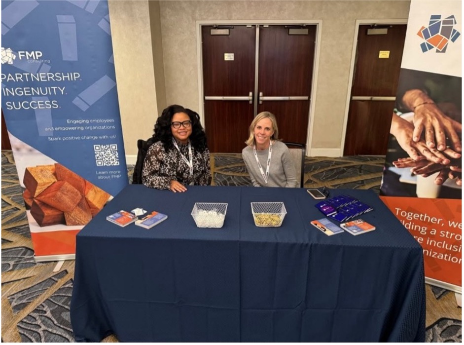 Photograph of two women sitting behind a table covered with a dark blue cloth at a conference. 