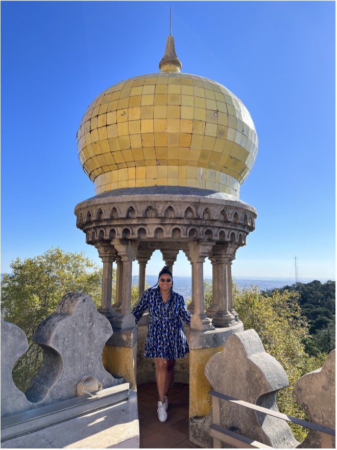 Photograph of a person standing inside a small, ornate stone pavilion topped with a large golden dome under a clear blue sky. Surrounding the pavilion are decorative stone railings and lush green trees.