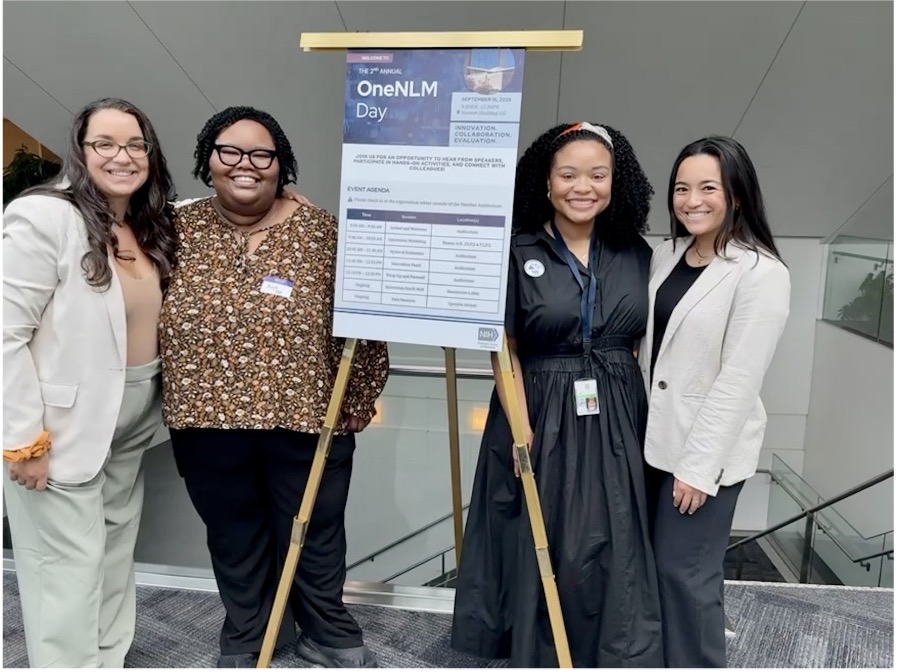 Photograph of four people standing beside a tripod easel holding a schedule board for OneNLM Day.