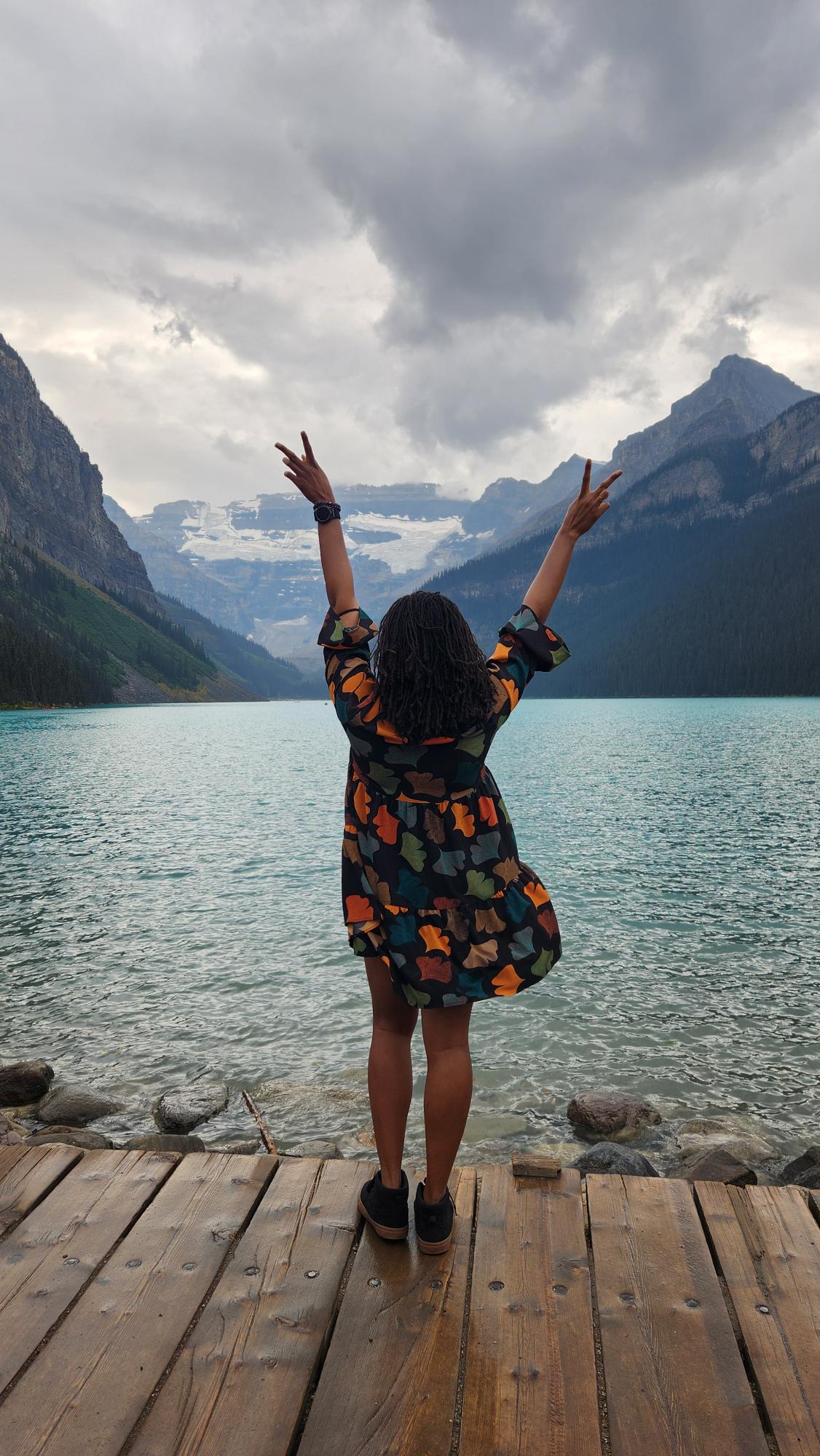 A woman with her back to the camera and holding her hands in peace signs, standing in front of a lake surrounded by mountains. 