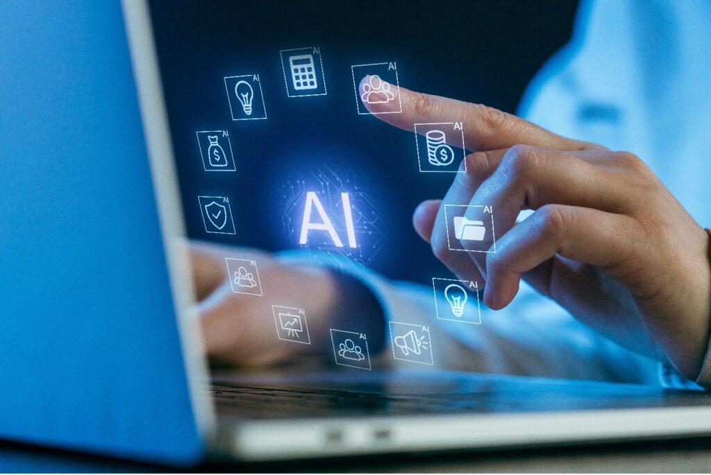 Photograph showing a person interacting with a laptop displaying glowing AI-related icon in the middle of the screen, surrounded by other icons, including a lightbulb, calculator, user profiles, and a dollar sign. 