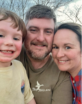 Three people, one child and two adults, smile outdoors with leafless trees and cloudy sky in background.