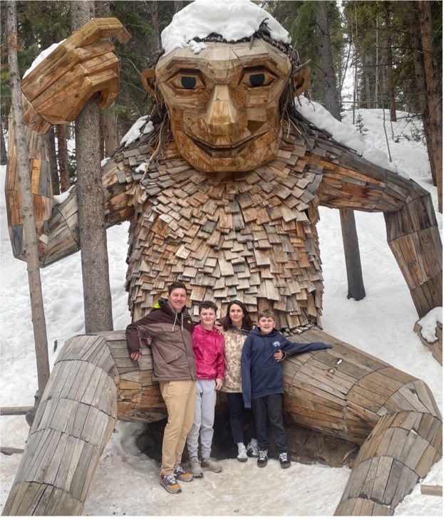 A family standing in front of a large figure made of wood. A snow covered forest is in the background.