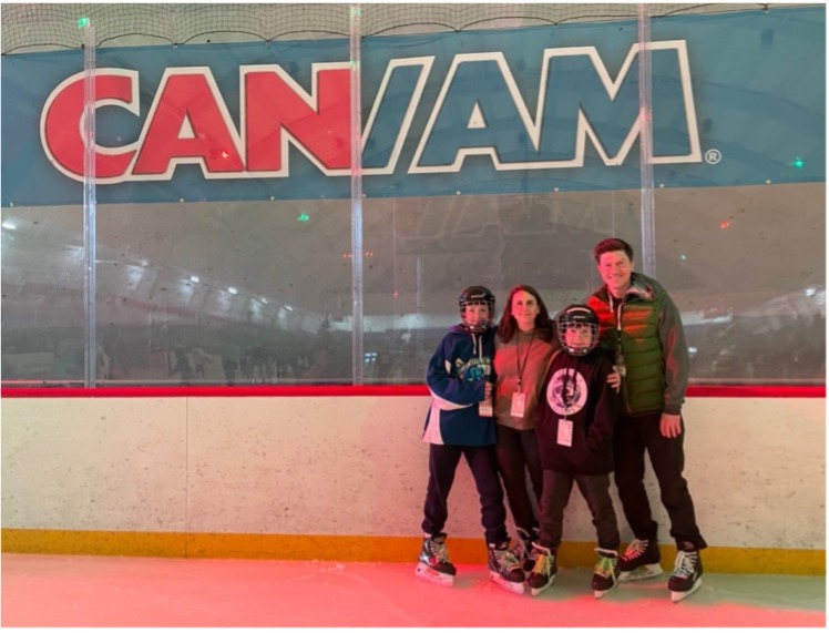 A family in ice skates standing in a group on an ice rink. A sign reading CAN/AM is behind them.