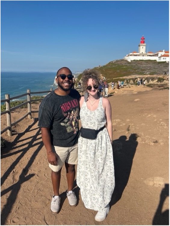 Two people standing by the ocean and in front of a light house in the distance. 