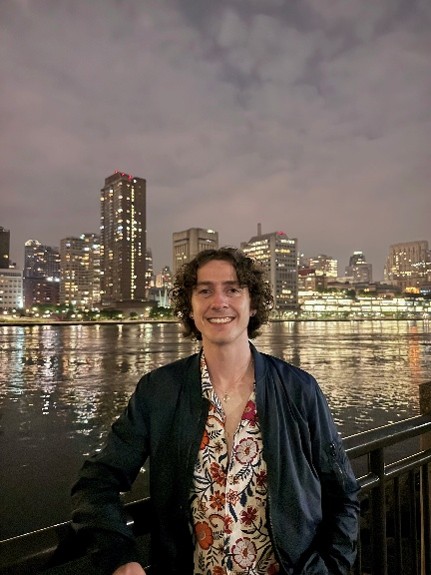 A man standing at night in front of a river with city buildings beyond the river.