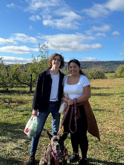 A man and woman standing in a field with a dog, holding bags of apples.