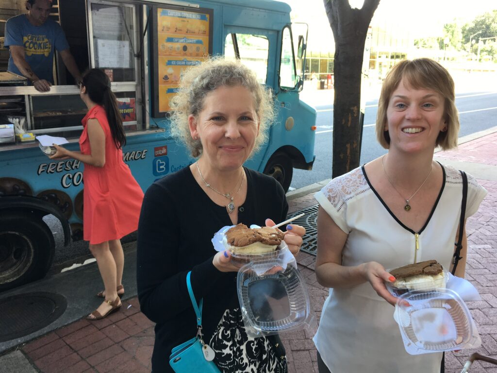 Two people standing in front of a food truck, holding containers of food and smiling.